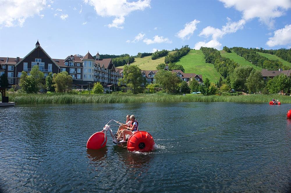 Two riders on a red water bike on the Mill Pond, with green ski runs behind