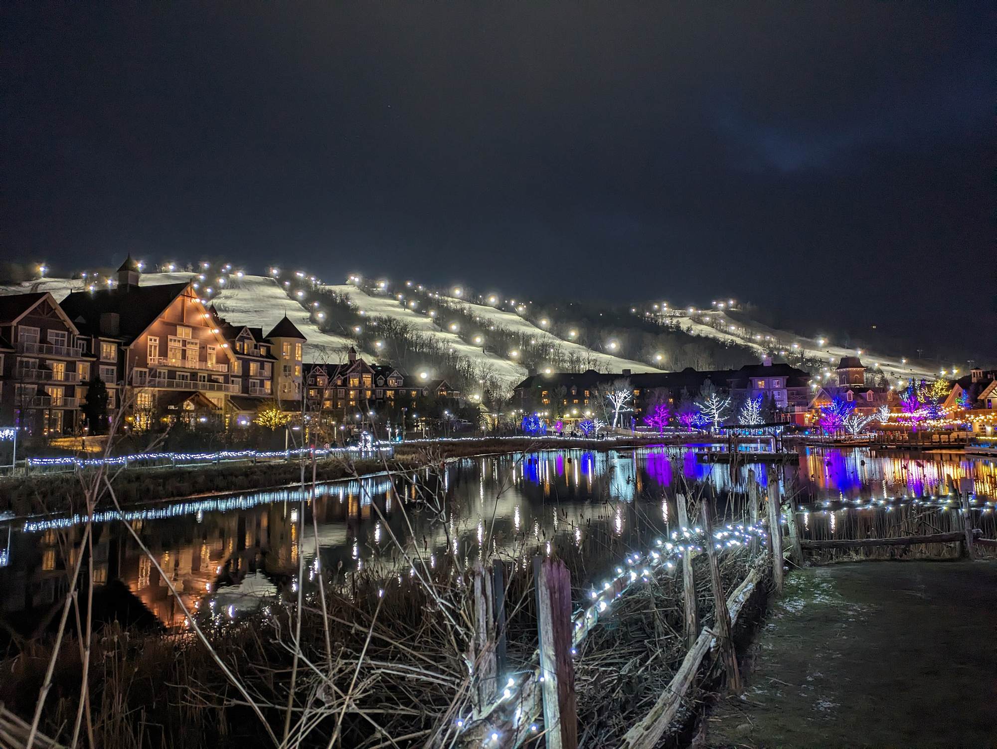 Blue Mountain Village at night, its lodges and trees lit up and reflected in the pond, with floodlit ski runs on the hill behind