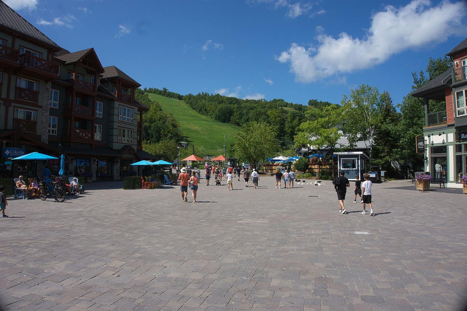 The Village plaza in August with people strolling, patio umbrellas and the green ski hill behind
