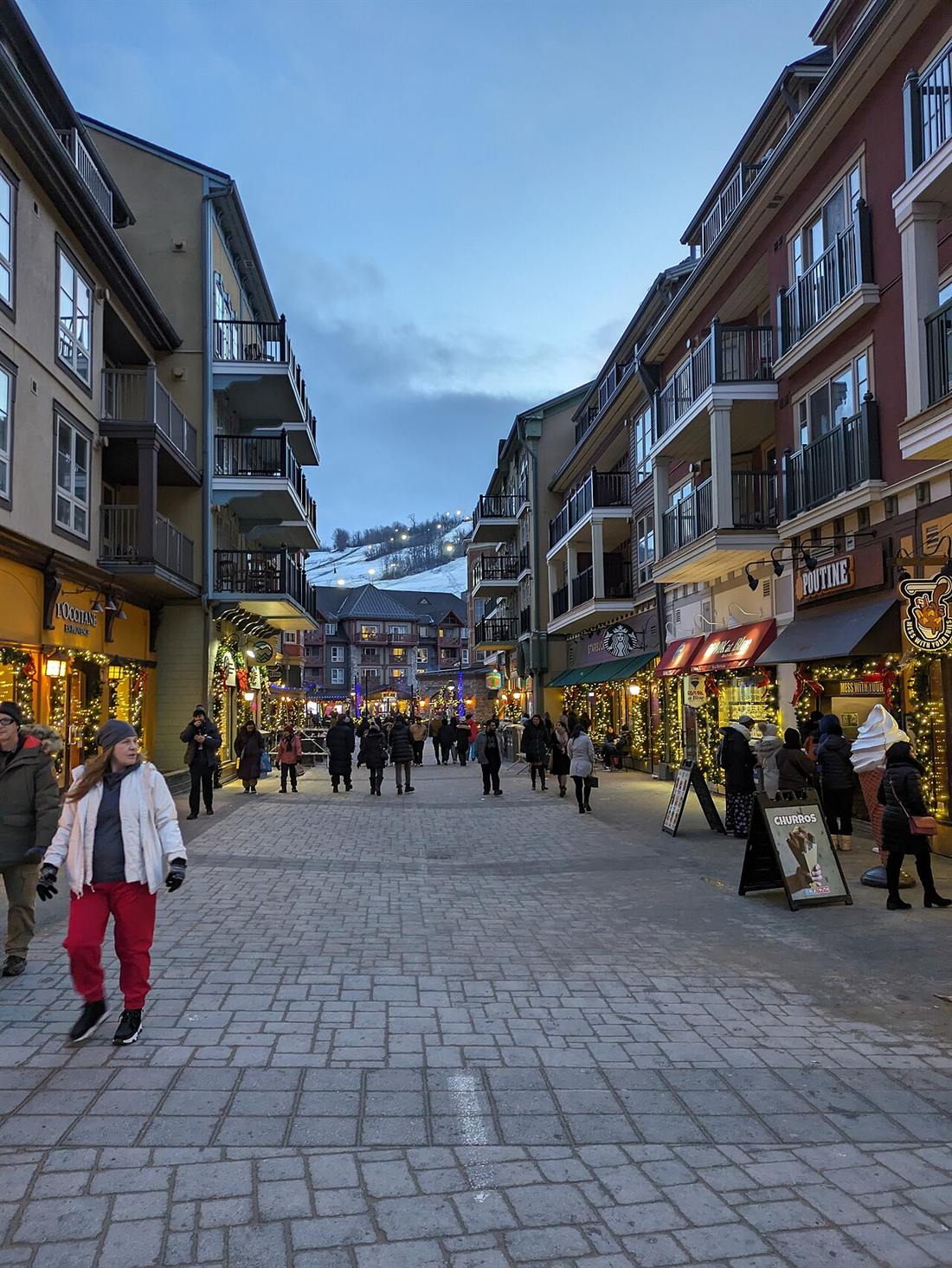 A pedestrian street in Blue Mountain Village at dusk in January, shops lit up and the ski hill at the end of the street